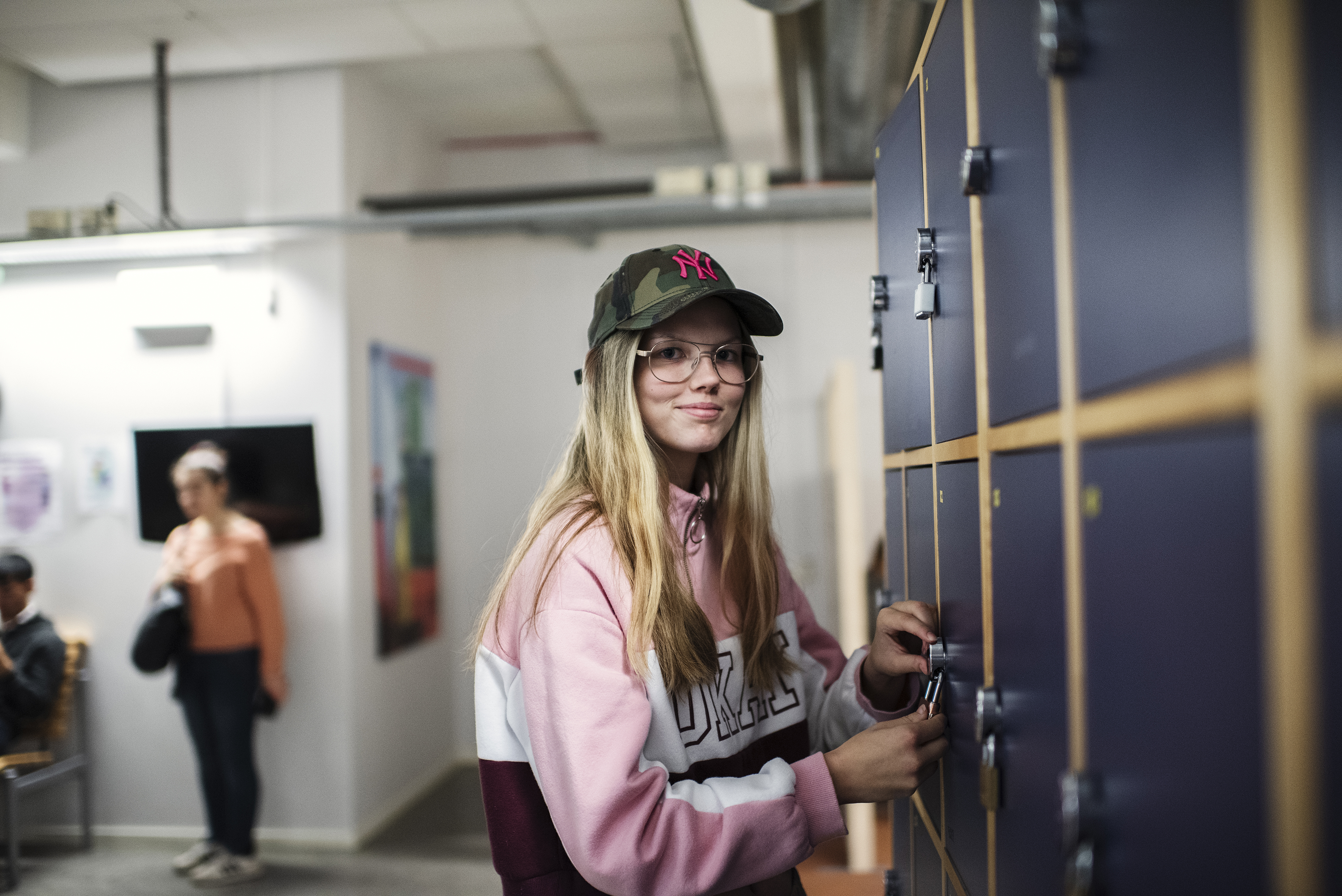 En gymnasieelev öppnar ett skåp i korridoren. Eleven bär glasögon och keps, och ser mot kameran med ett leende.
