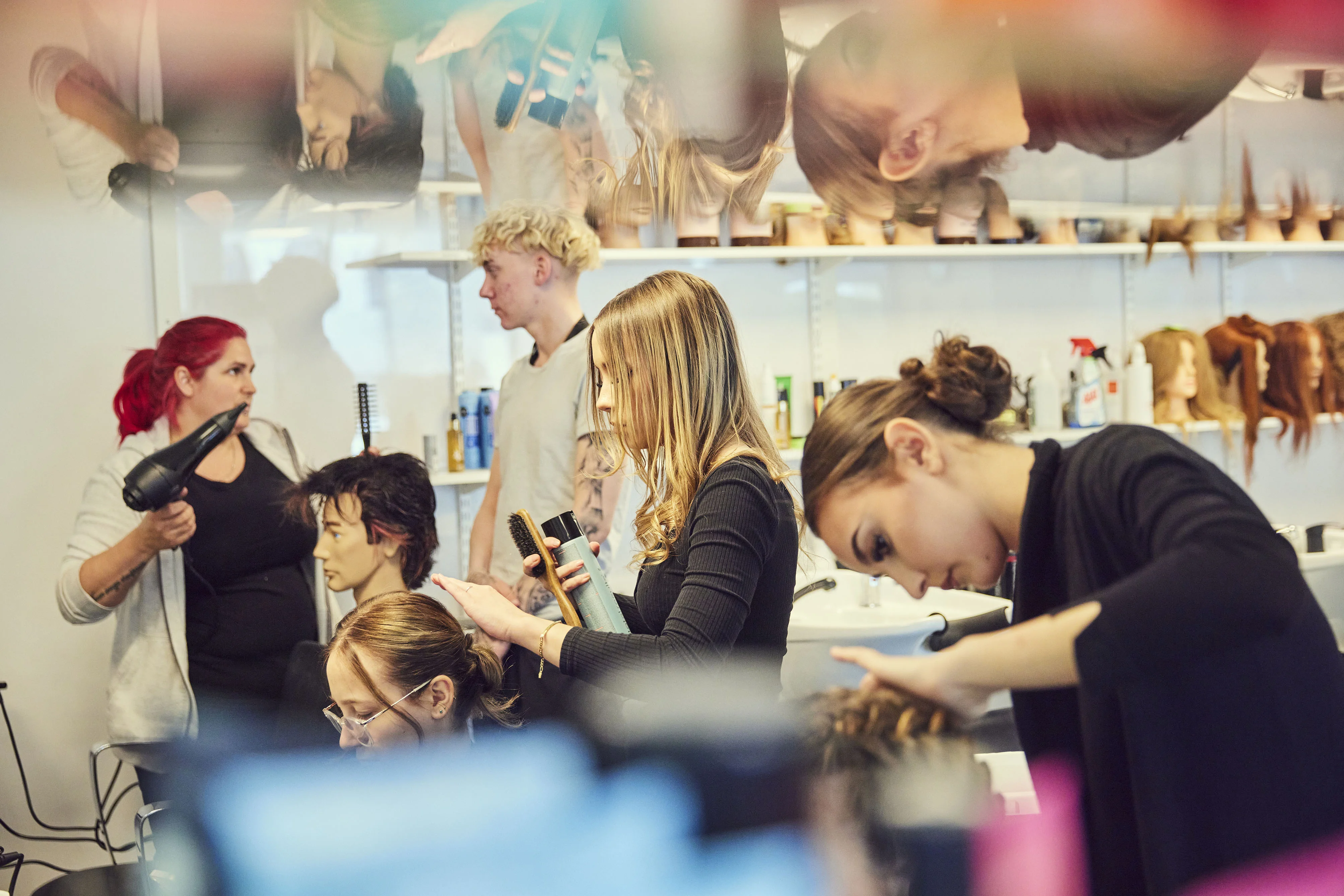 Elever tränar på hårstyling i salongmiljö, omgivna av speglar, frisördockor och hårprodukter.
