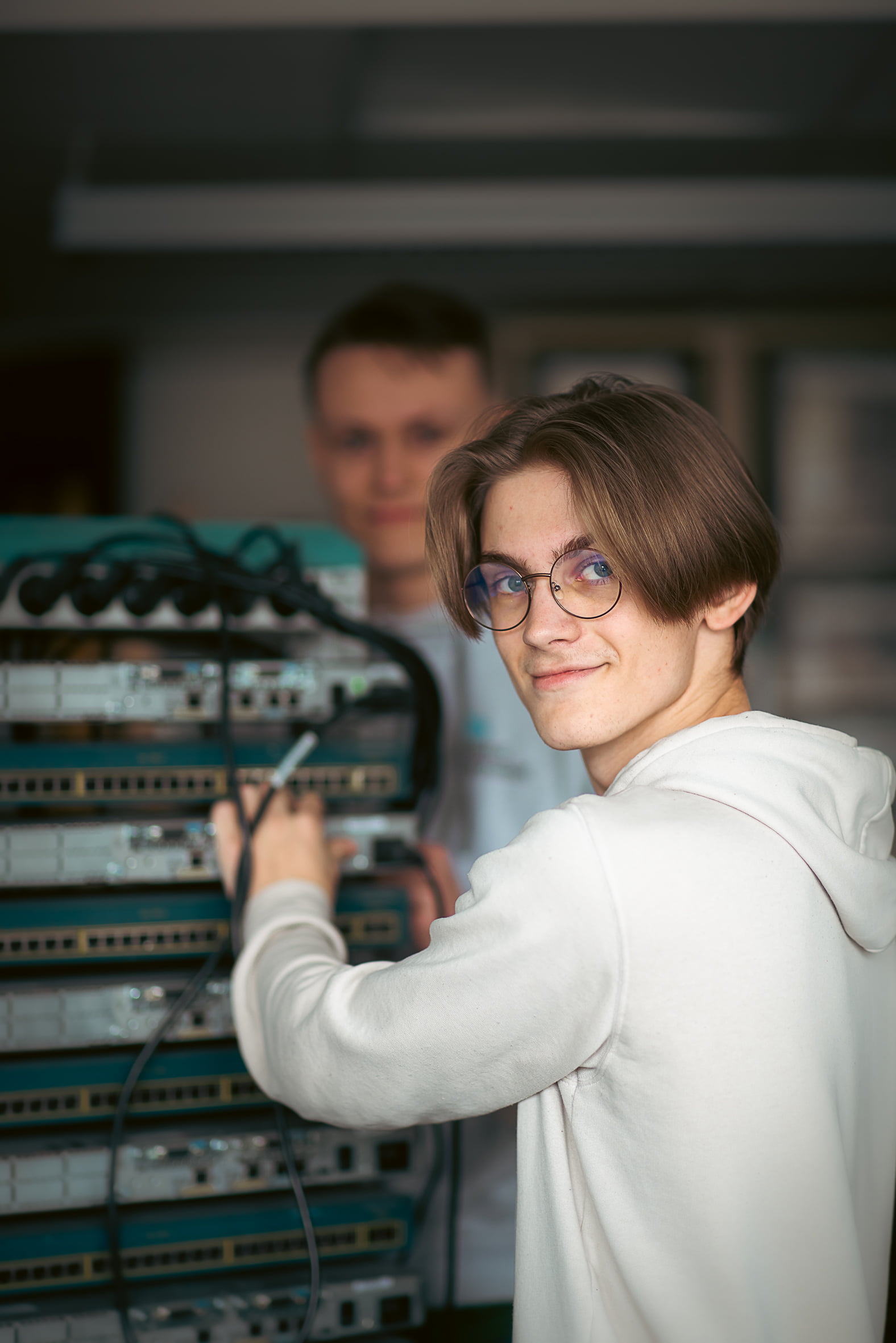 Elev kopplar nätverkskablar i en serverhall och ler mot kameran.