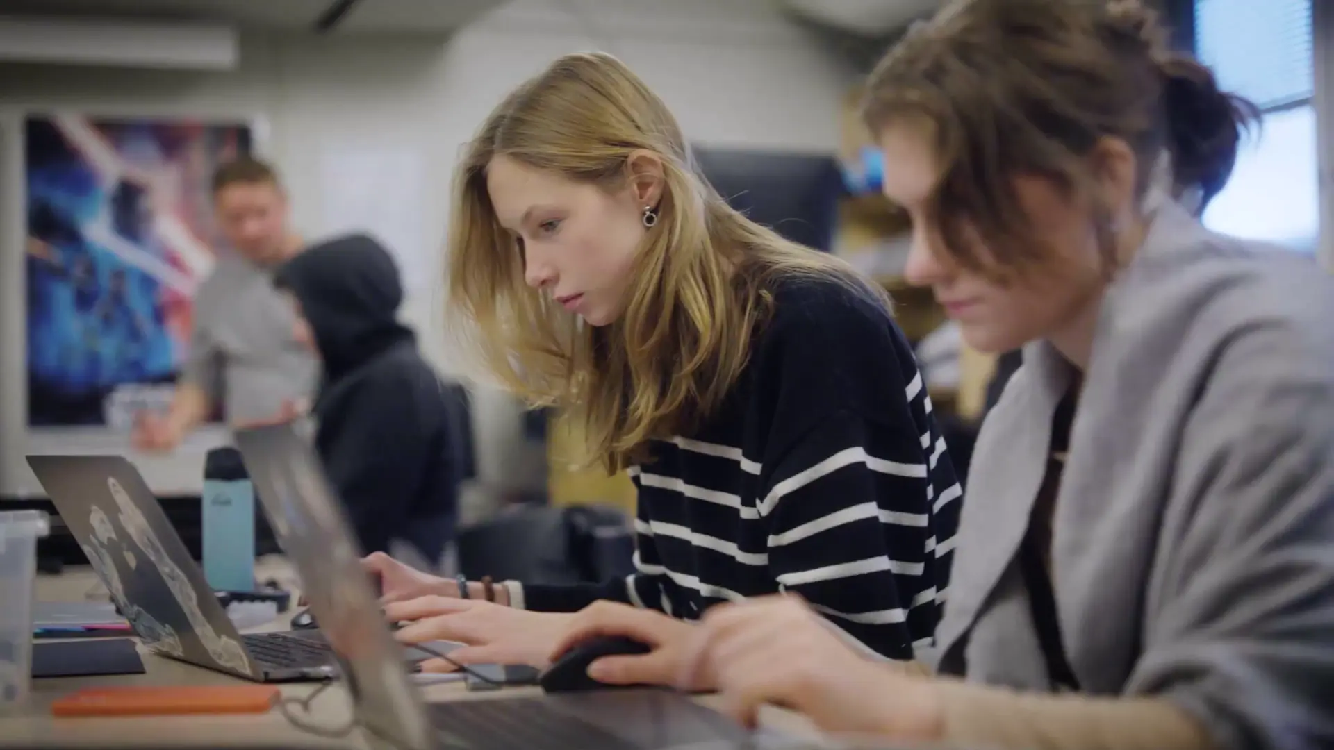 Två elever sitter vid ett långbord och arbetar koncentrerat vid sina bärbara datorer i en datasal eller ett kreativt klassrum.
