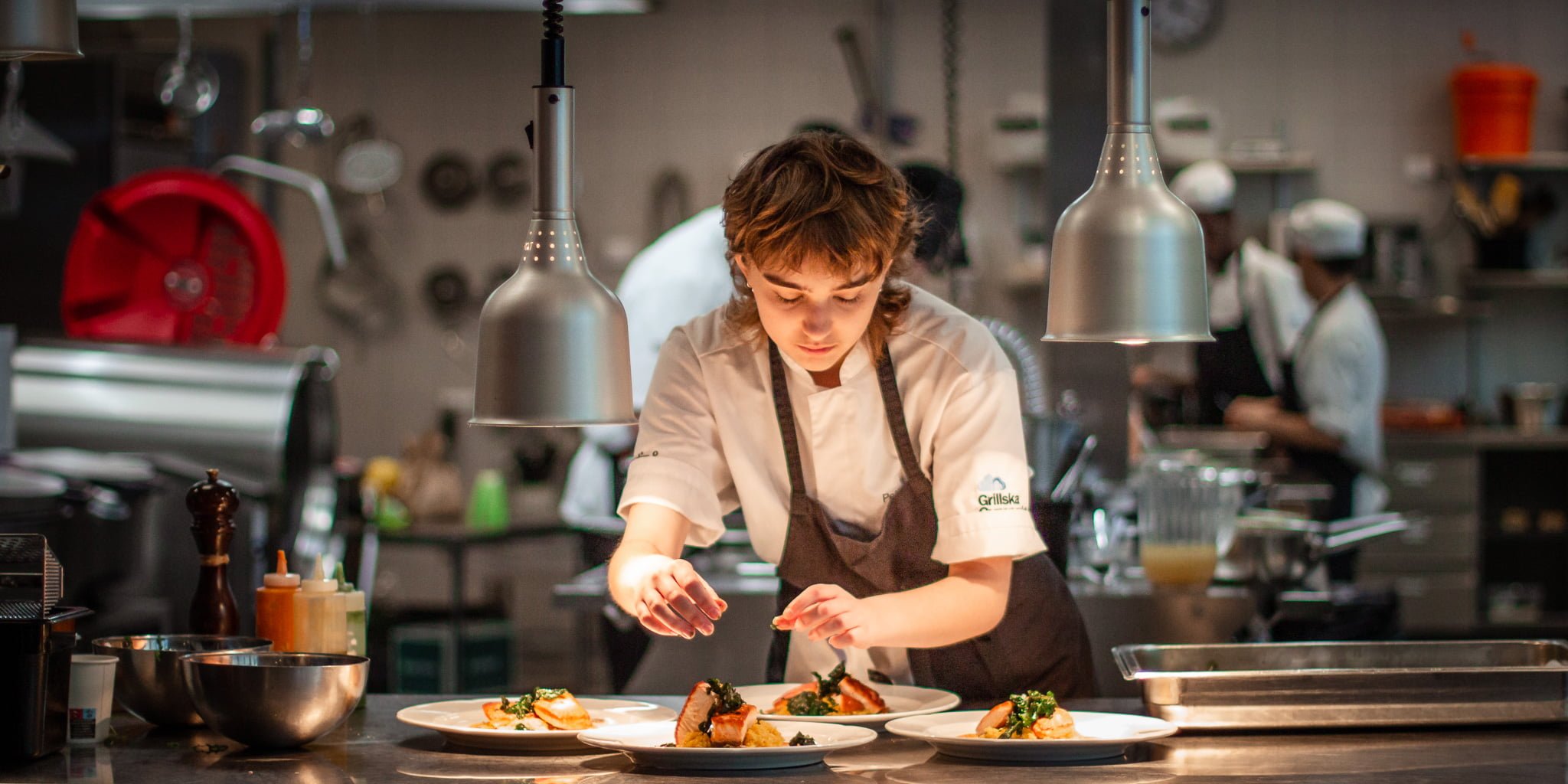 Elev i kockkläder lägger upp mat på tallrikar i ett restaurangkök.