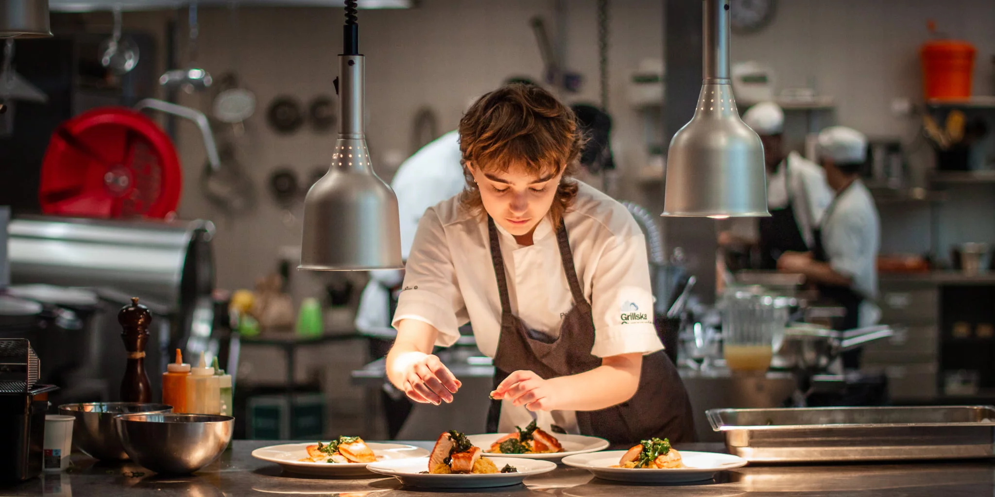 Elev i kockkläder lägger upp mat på tallrikar i ett restaurangkök.