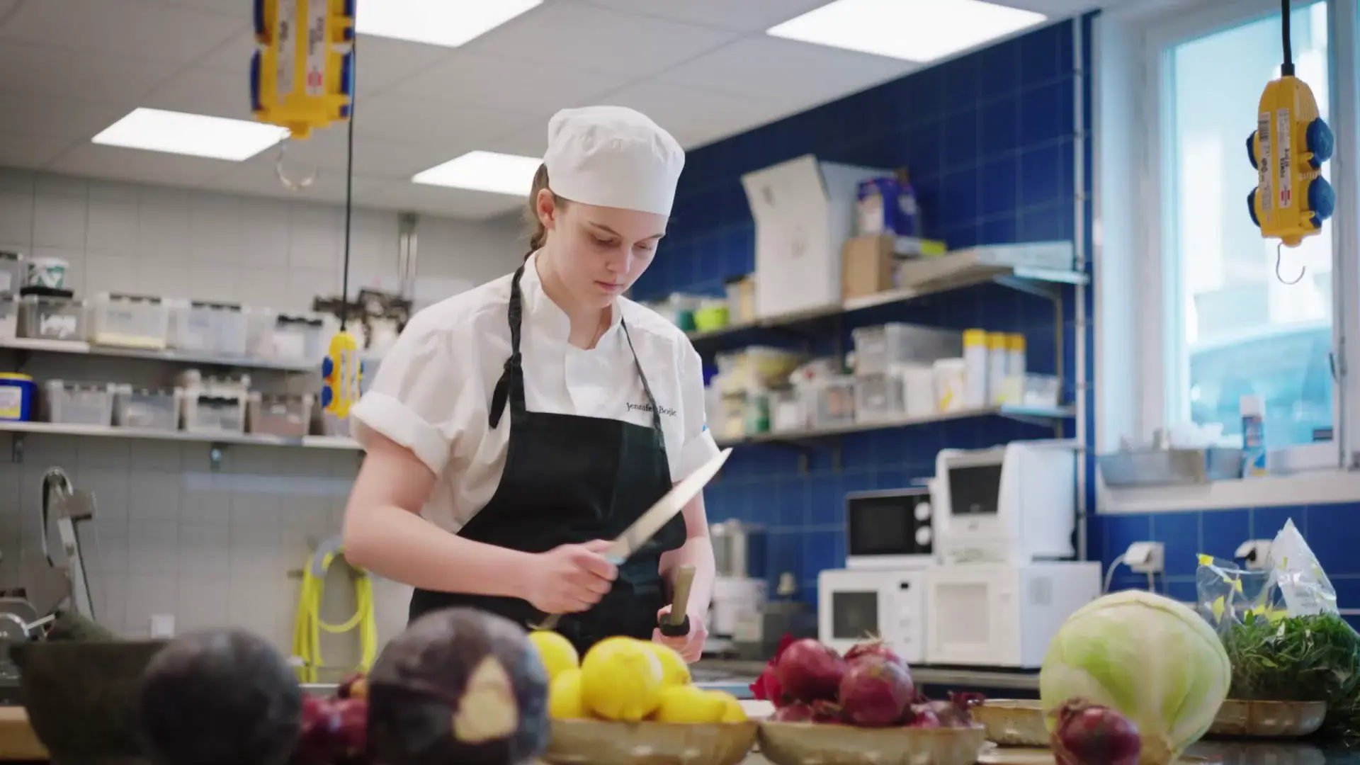 En elev på restaurangprogrammet, klädd i kockmössa och förkläde, förbereder grönsaker i ett storkök. På bänken framför syns kålhuvuden, citroner och rödlök.