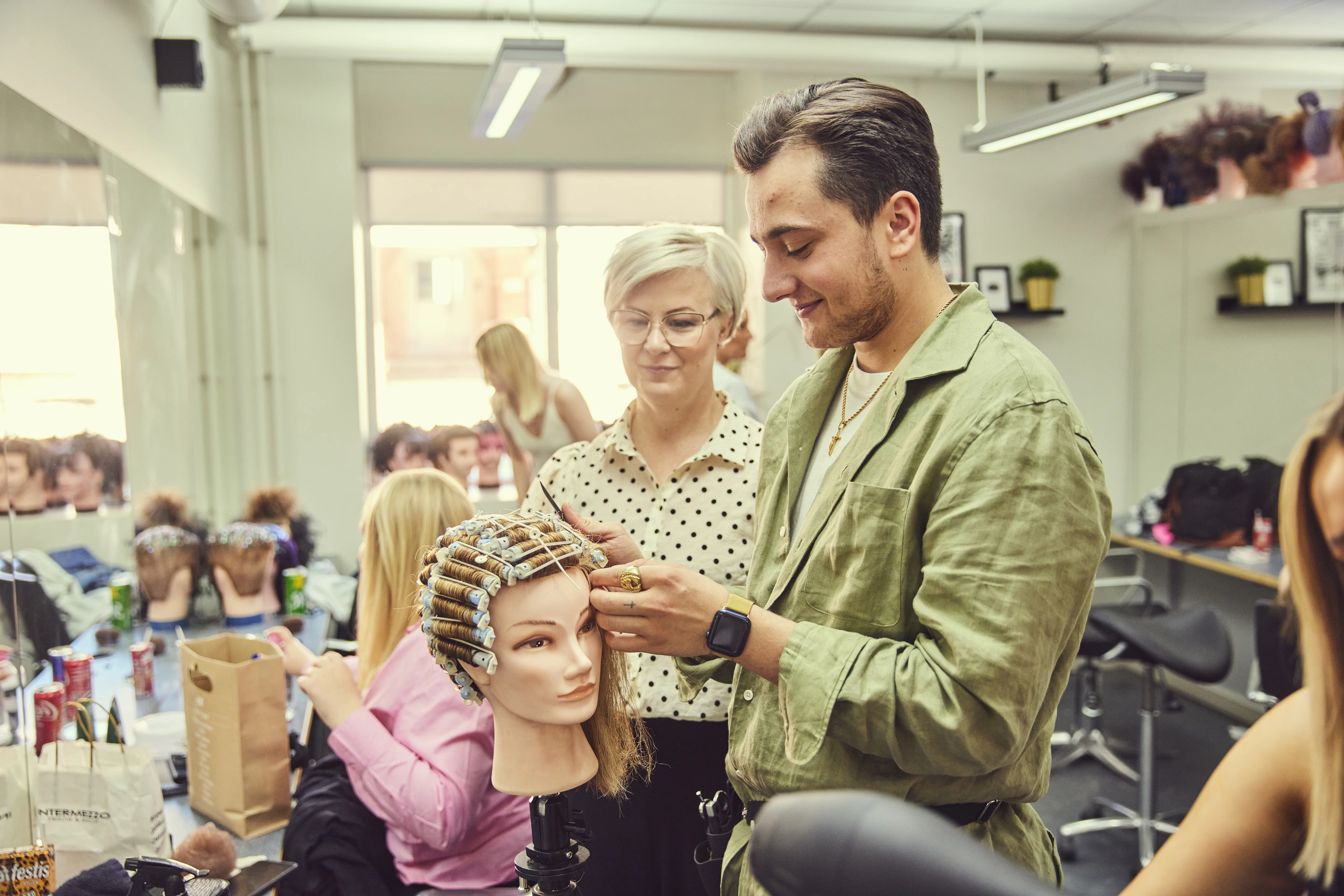 Elev på frisörutbildning tränar permanentteknik på en övningsdocka under handledning av en lärare. 