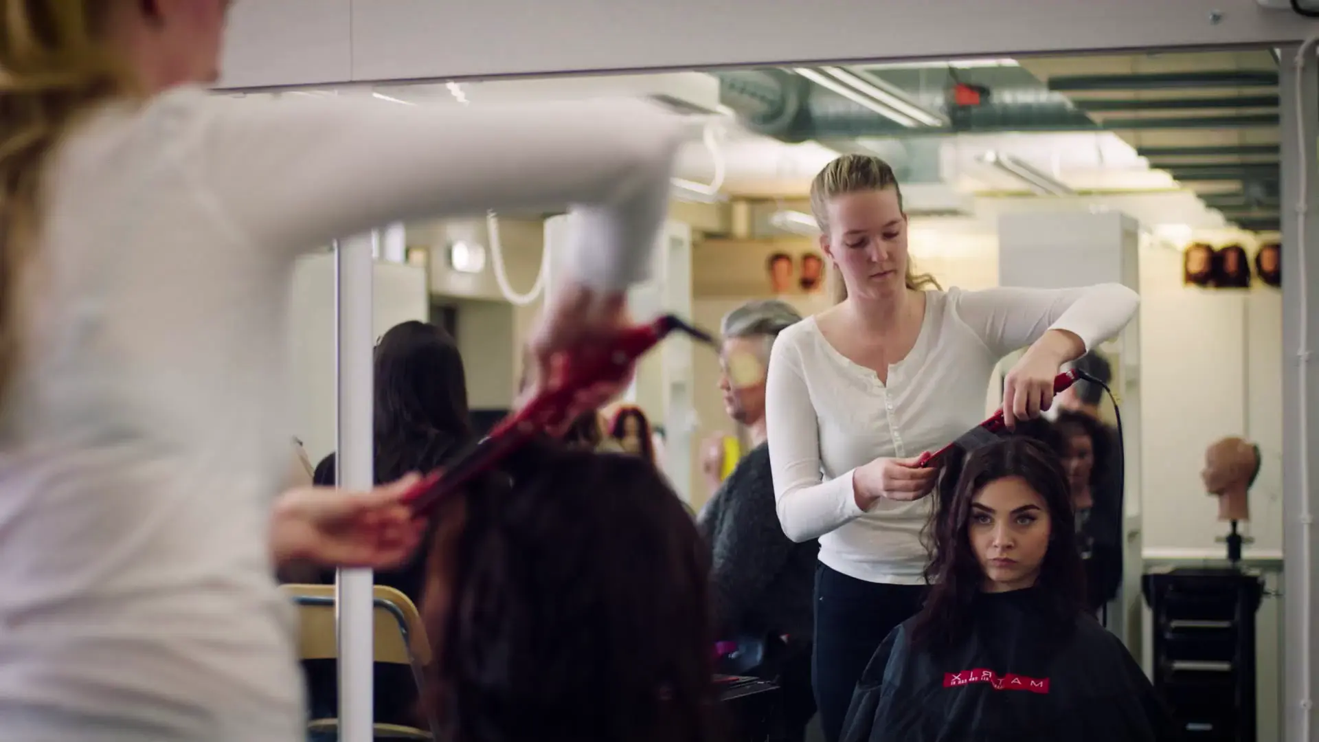 Elever på frisörprogrammet övar styling i en salongsmiljö. En elev står upp och använder en locktång på en annan elev som sitter framför en spegel. I bakgrunden syns frisörhuvuden för övning.