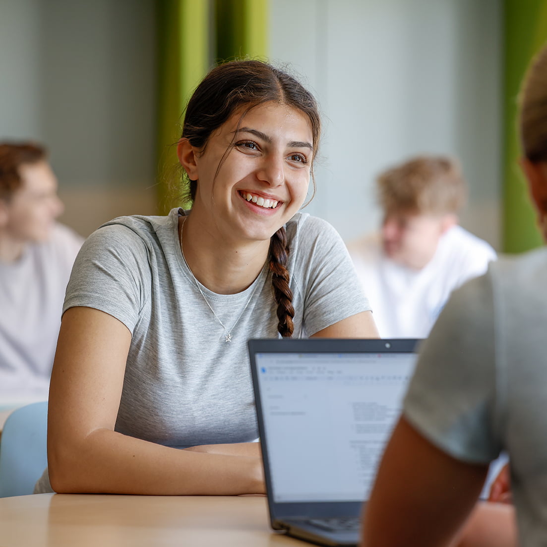 En tjej i grå t-shirt ler och pratar med en annan elev. En laptop står på bordet. Klassrumsmiljö med ljusa färger.
