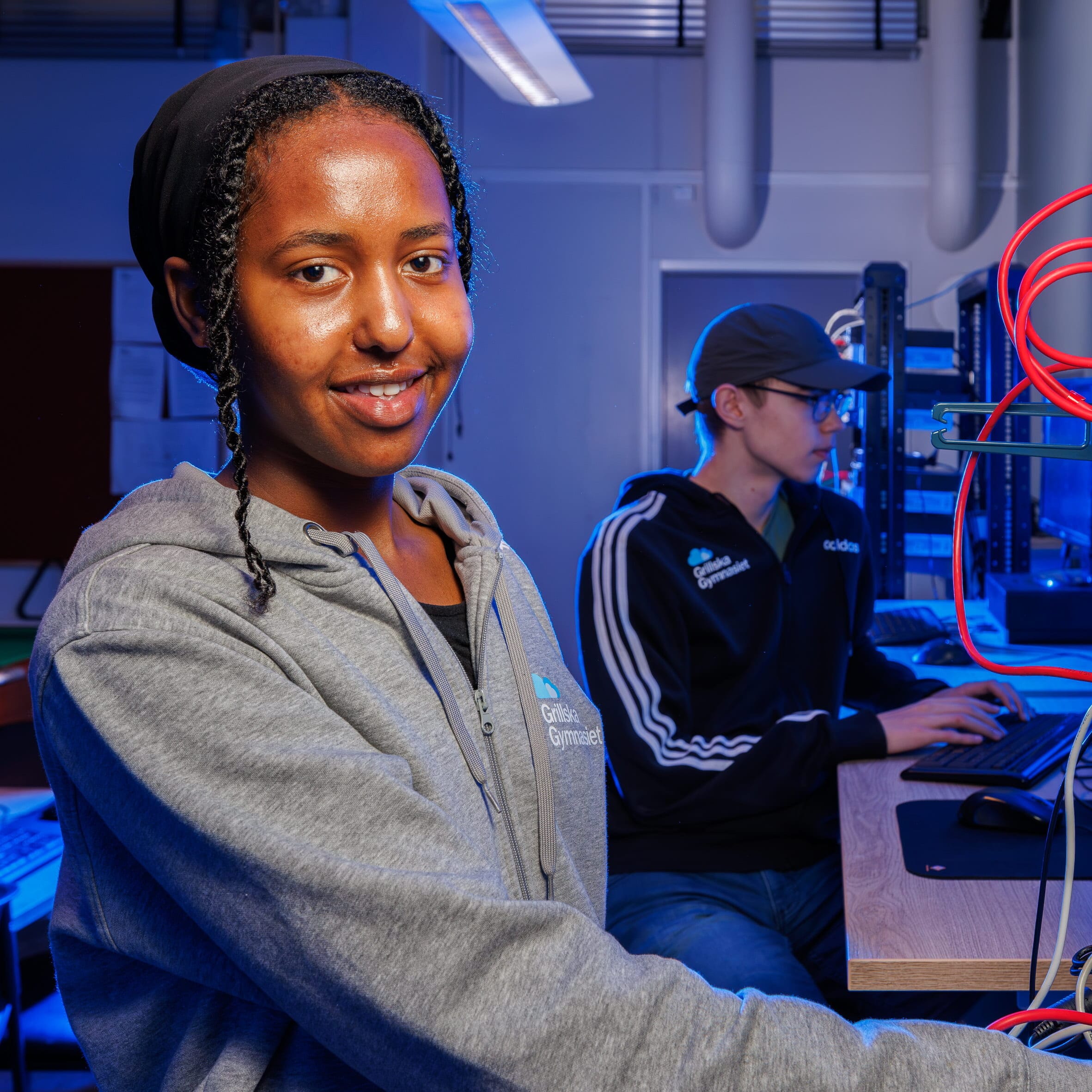Två elever arbetar vid nätverksutrustning. En tjej tittar mot kameran och ler, en kille fokuserar på en dator. Blå belysning och teknisk miljö.