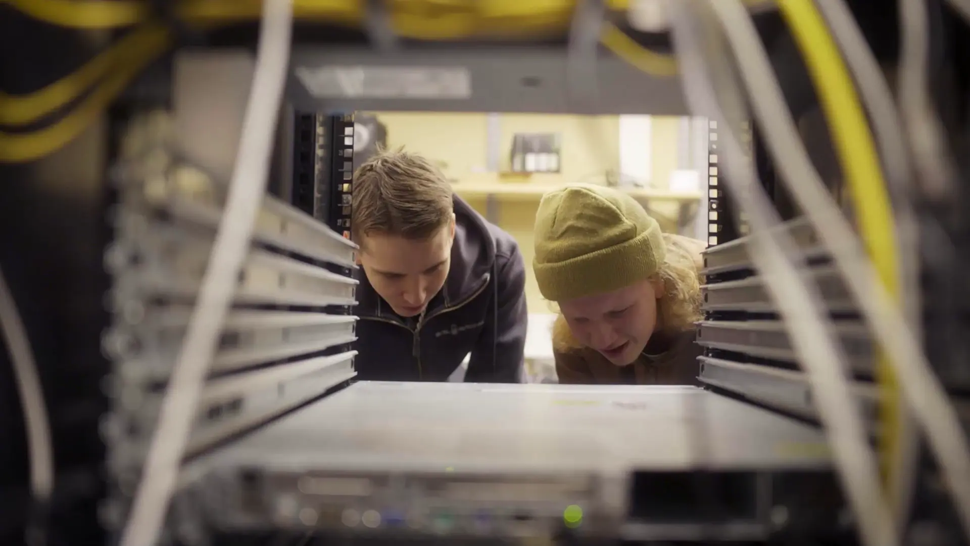 Två elever böjer sig fram och undersöker teknisk utrustning inuti ett serverrack. Bilden är tagen inifrån racket, vilket ramar in eleverna med nätverkskablar och hårdvara.