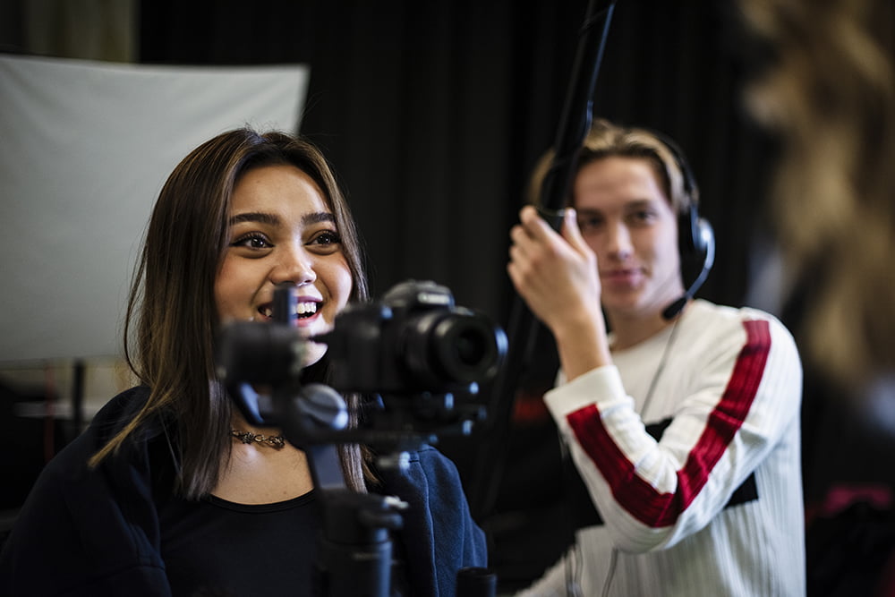 Två glada elever från samhällsvetenskaps&shy;programmet media programmet. En av dem filmar och den andra eleven håller en kamera mikrofon.