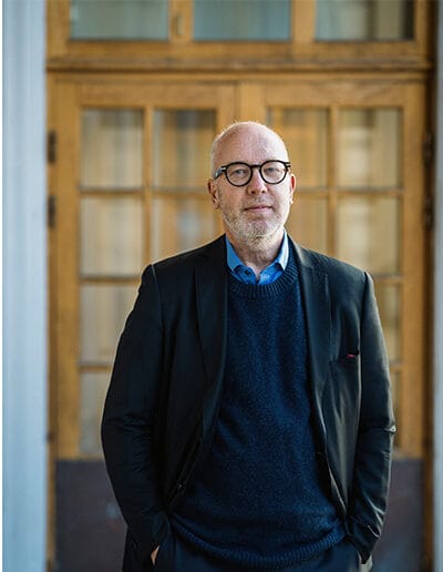 En man, Patriq Fagerstedt, med glasögon står framför en skolbyggnads entré. Bakom honom syns trädörrar med glasrutor.