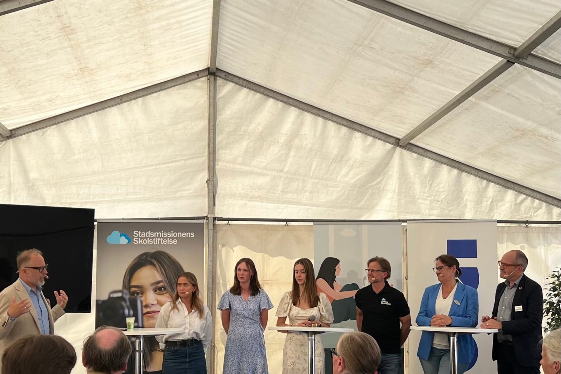 Ett panelsamtal med sju vuxna personer under ett tält. Bakom syns skyltar från skolstiftelse och andra organisationer.