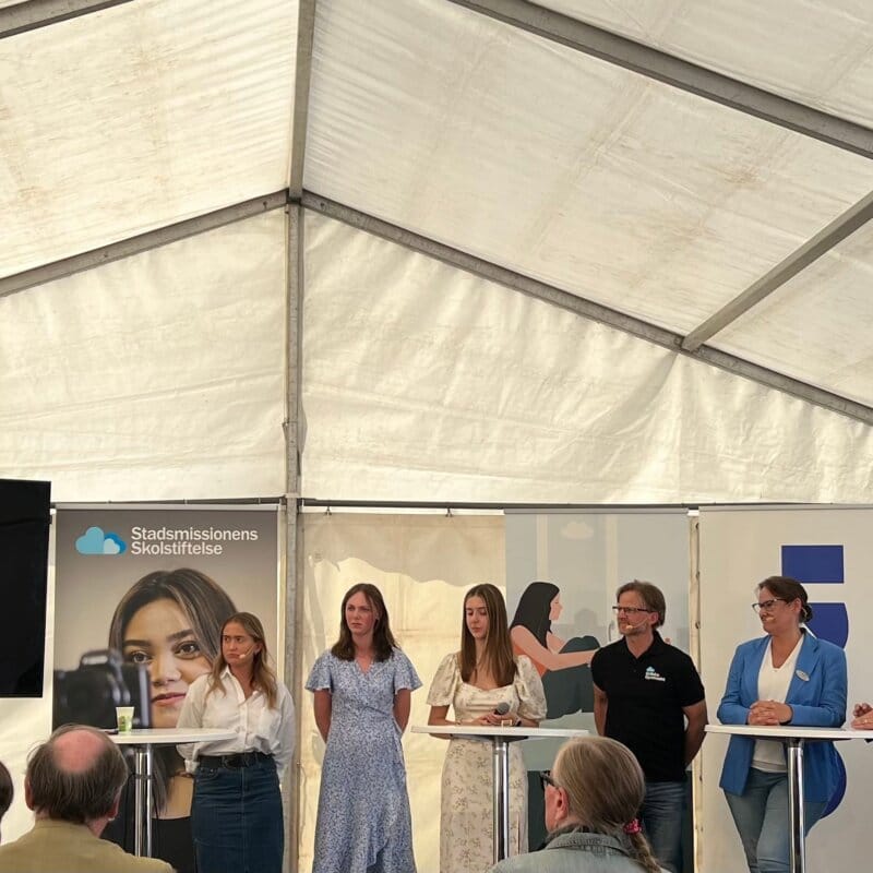 Ett panelsamtal med sju vuxna personer under ett tält. Bakom syns skyltar från skolstiftelse och andra organisationer.