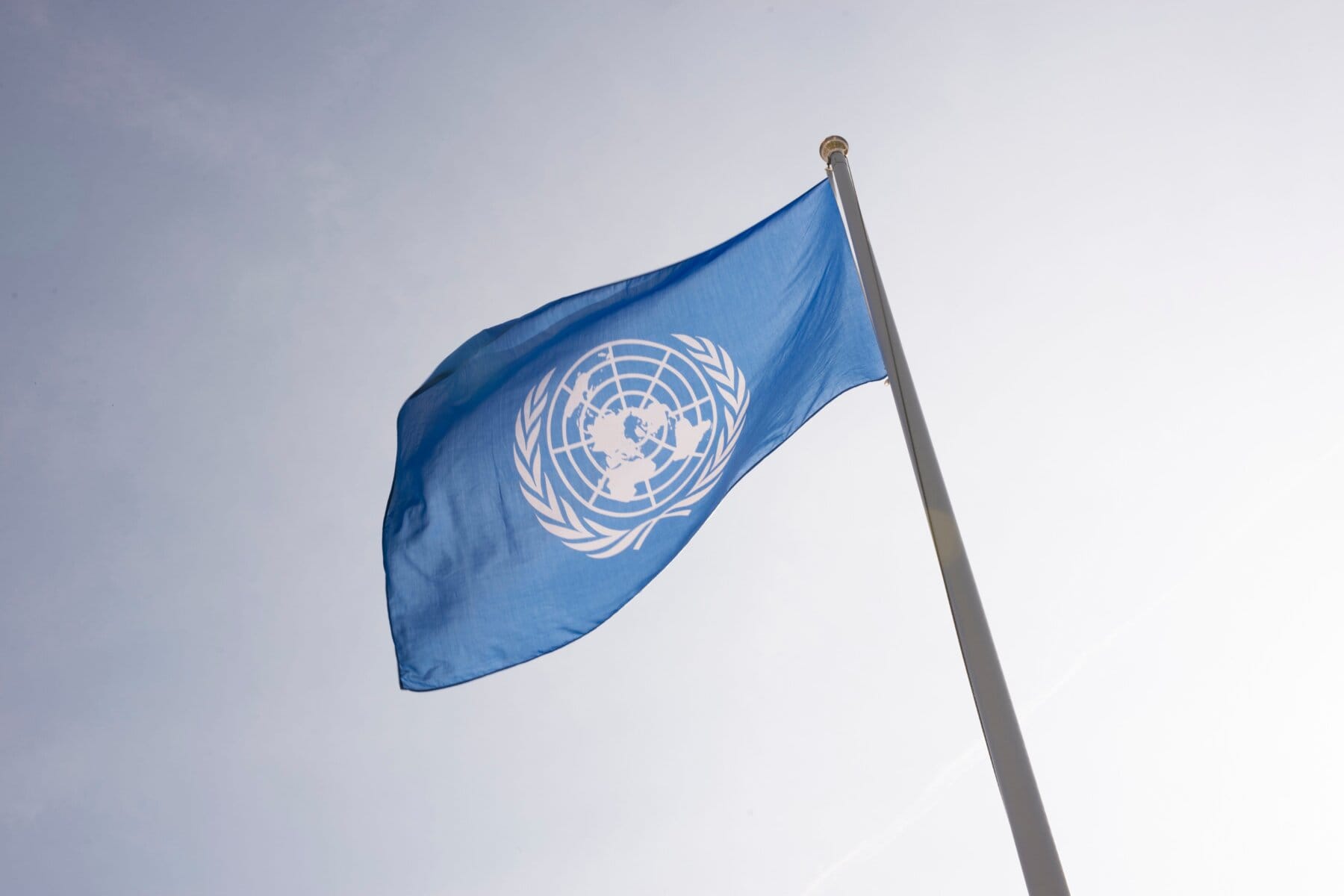 En blå flagga med FN:s symbol vajar på en flaggstång mot ljus himmel.