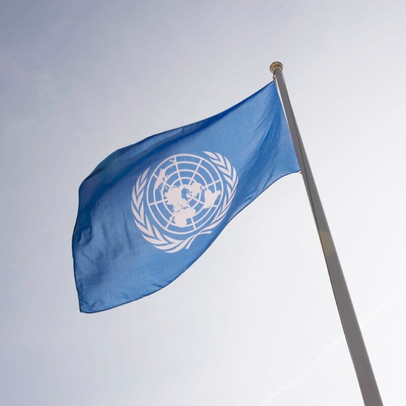 En blå flagga med FN:s symbol vajar på en flaggstång mot ljus himmel.