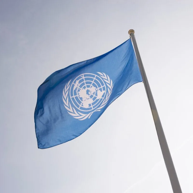 En blå flagga med FN:s symbol vajar på en flaggstång mot ljus himmel.