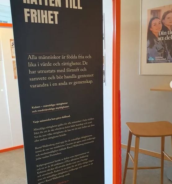 En svart informationsskylt med vit text om allas rätt till frihet. Texten står i en skolmiljö, intill en affisch.
